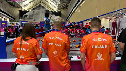 Trzech uczestników polskiej drużyny, widocznych od tyłu, obserwuje zawody robotyczne. Mają pomarańczowe koszulki z napisem „POLAND” i logo FIRST Global. Przed nimi znajduje się arena z elementami konkursowymi, a w tle inne drużyny.