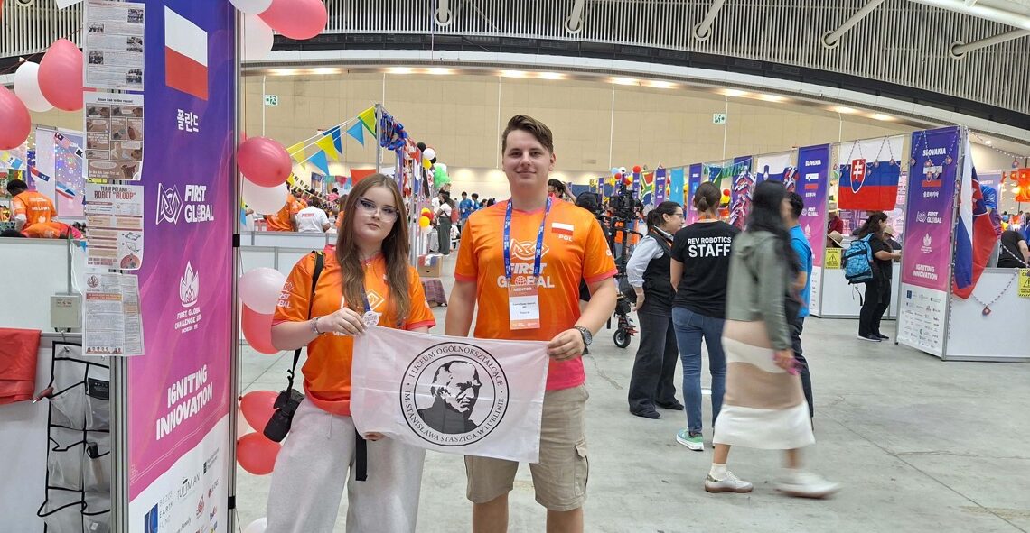 Dwoje młodych uczestników w pomarańczowych koszulkach FIRST Global trzyma białą flagę z godłem i nazwą I Liceum Ogólnokształcącego im. Stanisława Staszica w Lublinie. W tle widoczne są stoiska międzynarodowych drużyn, flagi i kolorowe dekoracje.