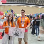 Dwoje młodych uczestników w pomarańczowych koszulkach FIRST Global trzyma białą flagę z godłem i nazwą I Liceum Ogólnokształcącego im. Stanisława Staszica w Lublinie. W tle widoczne są stoiska międzynarodowych drużyn, flagi i kolorowe dekoracje.