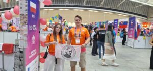 Dwoje młodych uczestników w pomarańczowych koszulkach FIRST Global trzyma białą flagę z godłem i nazwą I Liceum Ogólnokształcącego im. Stanisława Staszica w Lublinie. W tle widoczne są stoiska międzynarodowych drużyn, flagi i kolorowe dekoracje.