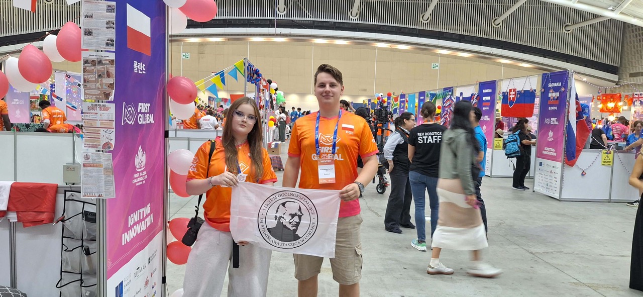 Dwoje młodych uczestników w pomarańczowych koszulkach FIRST Global trzyma białą flagę z godłem i nazwą I Liceum Ogólnokształcącego im. Stanisława Staszica w Lublinie. W tle widoczne są stoiska międzynarodowych drużyn, flagi i kolorowe dekoracje.