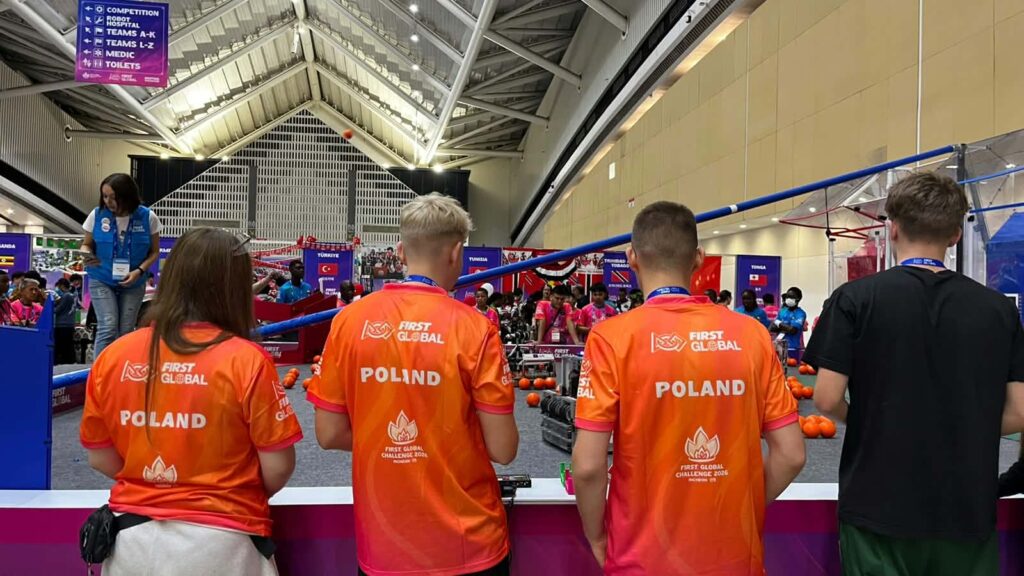 Czworo uczestników obserwuje zawody robotyczne FIRST Global. Troje z nich ma pomarańczowe koszulki z napisem „POLAND”. W tle znajduje się arena z robotami, pomarańczowymi piłkami i drużynami z różnych krajów.