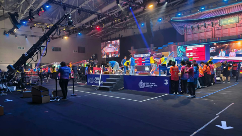Duża hala sportowa podczas międzynarodowych zawodów robotycznych FIRST Global. Na środku znajdują się stanowiska konkursowe, ekrany i kolorowe konstrukcje. Wokół stoją uczestnicy oraz organizatorzy. Halę oświetlają niebieskie i czerwone reflektory.