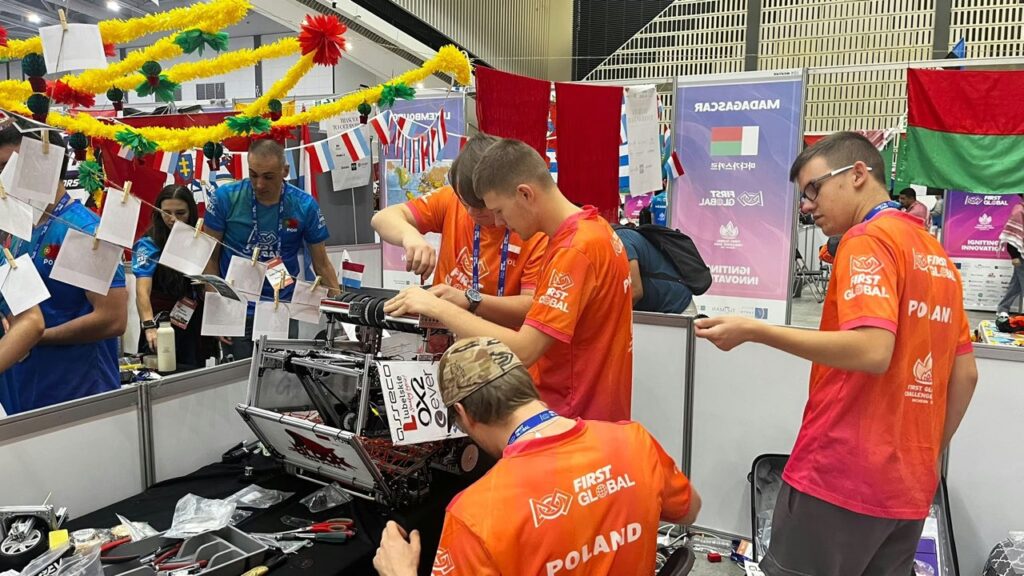 Członkowie polskiej drużyny FIRST Global naprawiają i regulują robota na stanowisku technicznym. Na stole leżą narzędzia i części konstrukcyjne. W tle widoczne są kolorowe dekoracje, flagi państw i uczestnicy innych drużyn.
