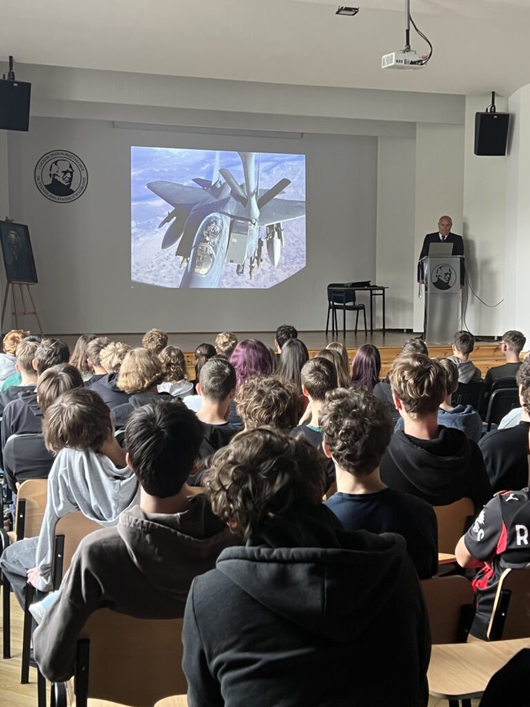 Prelegent przemawia do zgromadzonej publiczności na tle slajdu z ilustracją.