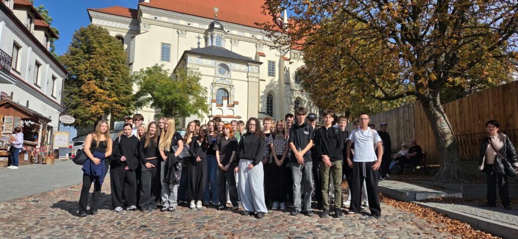 Uczniowie stoją na brukowanej uliczce przed okazałym, jasnym kościołem z czerwonym dachem i ozdobną fasadą. Po prawej stronie rośnie duże drzewo o jesiennych liściach, a po lewej widoczne są kamienice i stragany z pamiątkami.