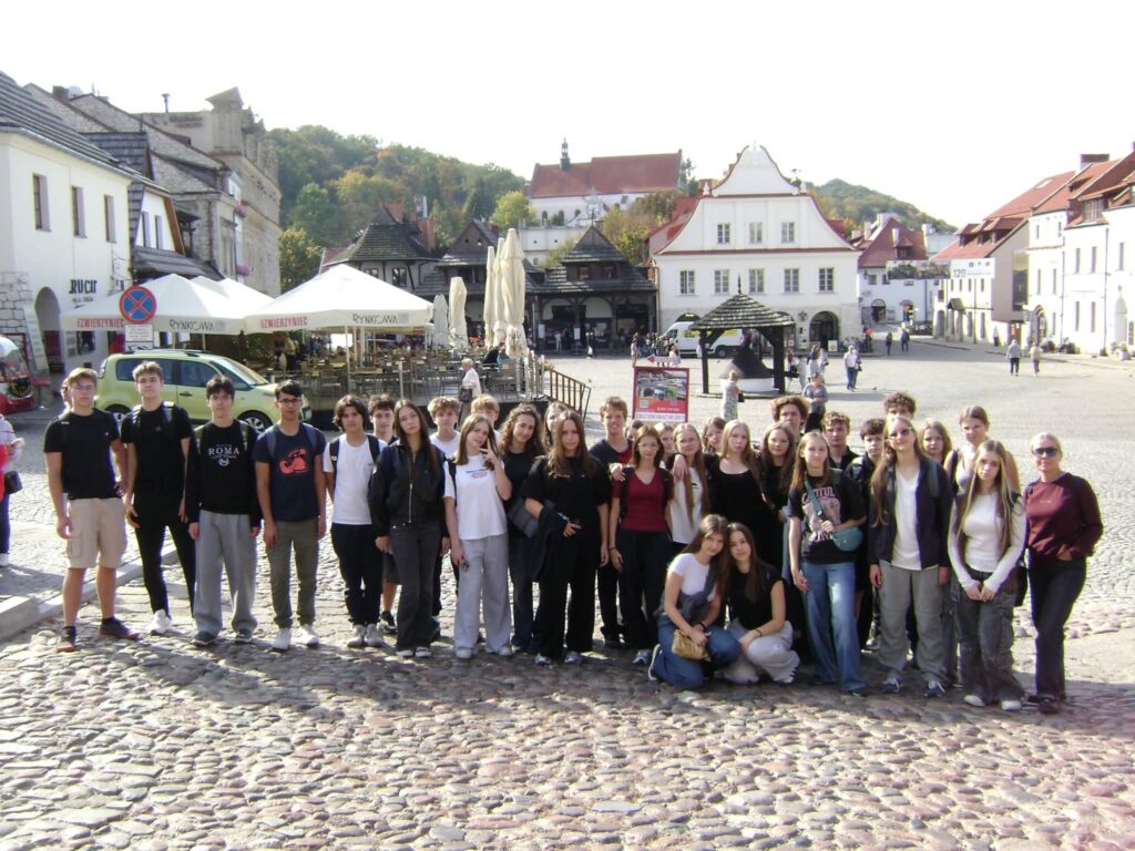 Duża grupa młodzieży wraz z opiekunką pozuje na brukowanym placu. W tle znajdują się zabytkowe kamienice, restauracyjne ogródki z parasolami oraz biały kościół. Zdjęcie wykonano w słoneczny dzień.