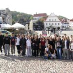 Duża grupa młodzieży wraz z opiekunką pozuje na brukowanym placu. W tle znajdują się zabytkowe kamienice, restauracyjne ogródki z parasolami oraz biały kościół. Zdjęcie wykonano w słoneczny dzień.