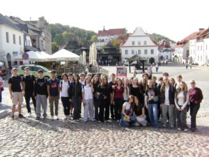 Duża grupa młodzieży wraz z opiekunką pozuje na brukowanym placu. W tle znajdują się zabytkowe kamienice, restauracyjne ogródki z parasolami oraz biały kościół. Zdjęcie wykonano w słoneczny dzień.