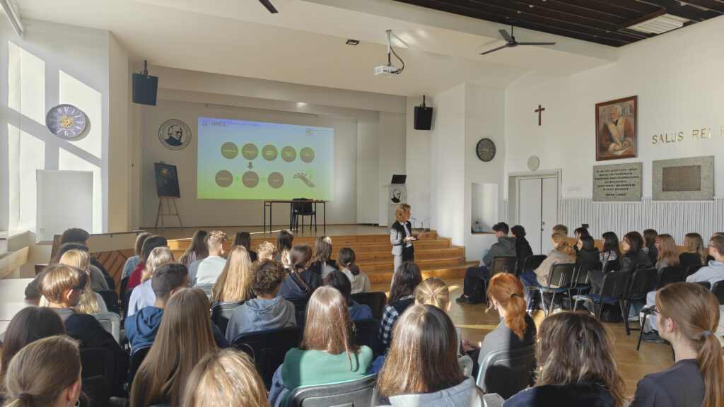 Widok od tyłu sali na uczniów uczestniczących w wykładzie. Przed drewnianą sceną stoi prelegentka w ciemnym garniturze, zwrócona w stronę młodzieży. Na ścianie za nią wyświetlany jest slajd z diagramem dotyczącym ekonomii. Po prawej stronie widoczne są portret, krzyż i tablice pamiątkowe.