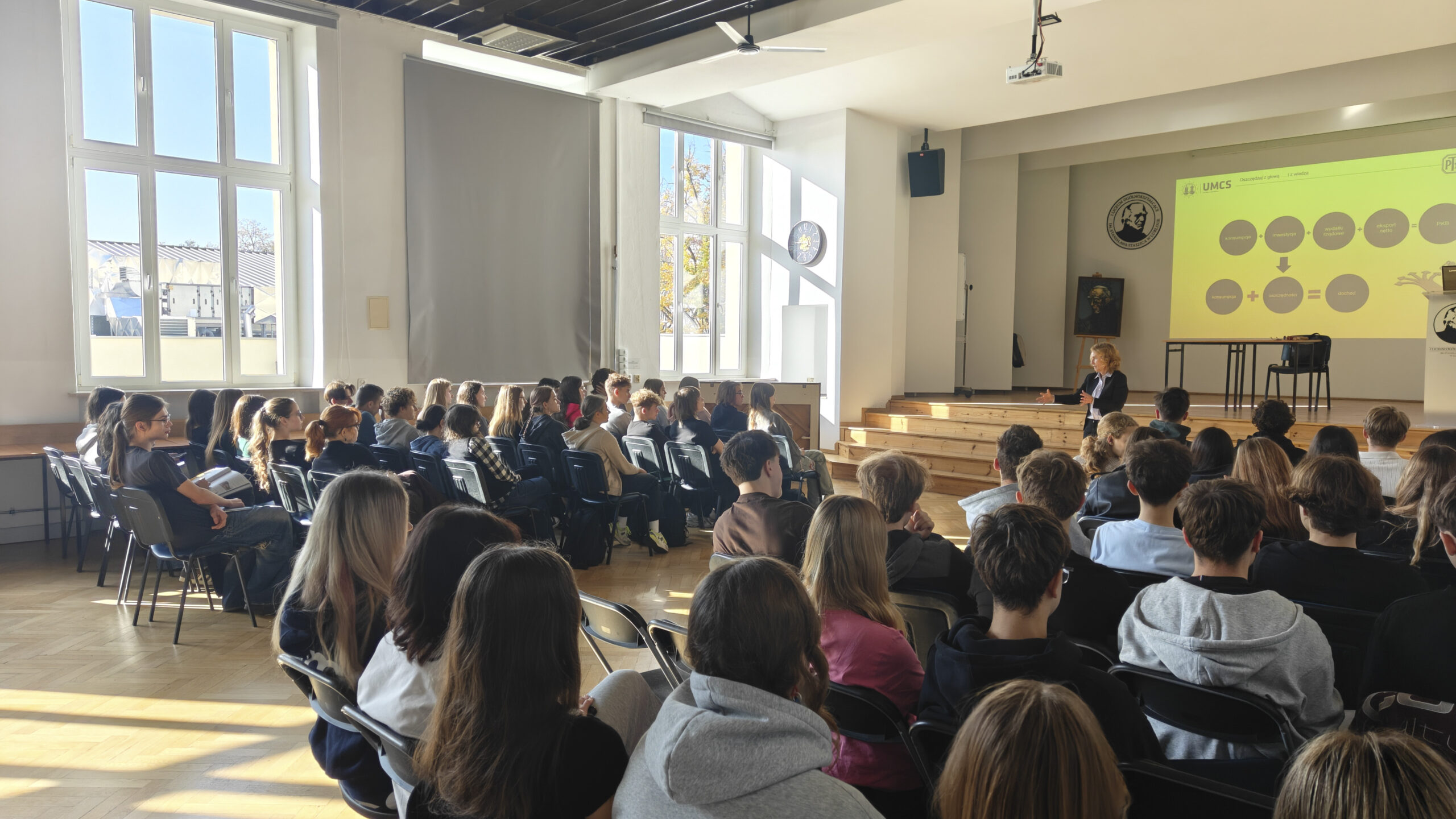 W jasnej auli szkolnej kobieta prowadzi wykład dla kilkudziesięciu uczniów siedzących w rzędach krzeseł. Na scenie wyświetlana jest prezentacja ekonomiczna z diagramami. Przez wysokie okna wpada światło słoneczne, oświetlając uczestników spotkania.